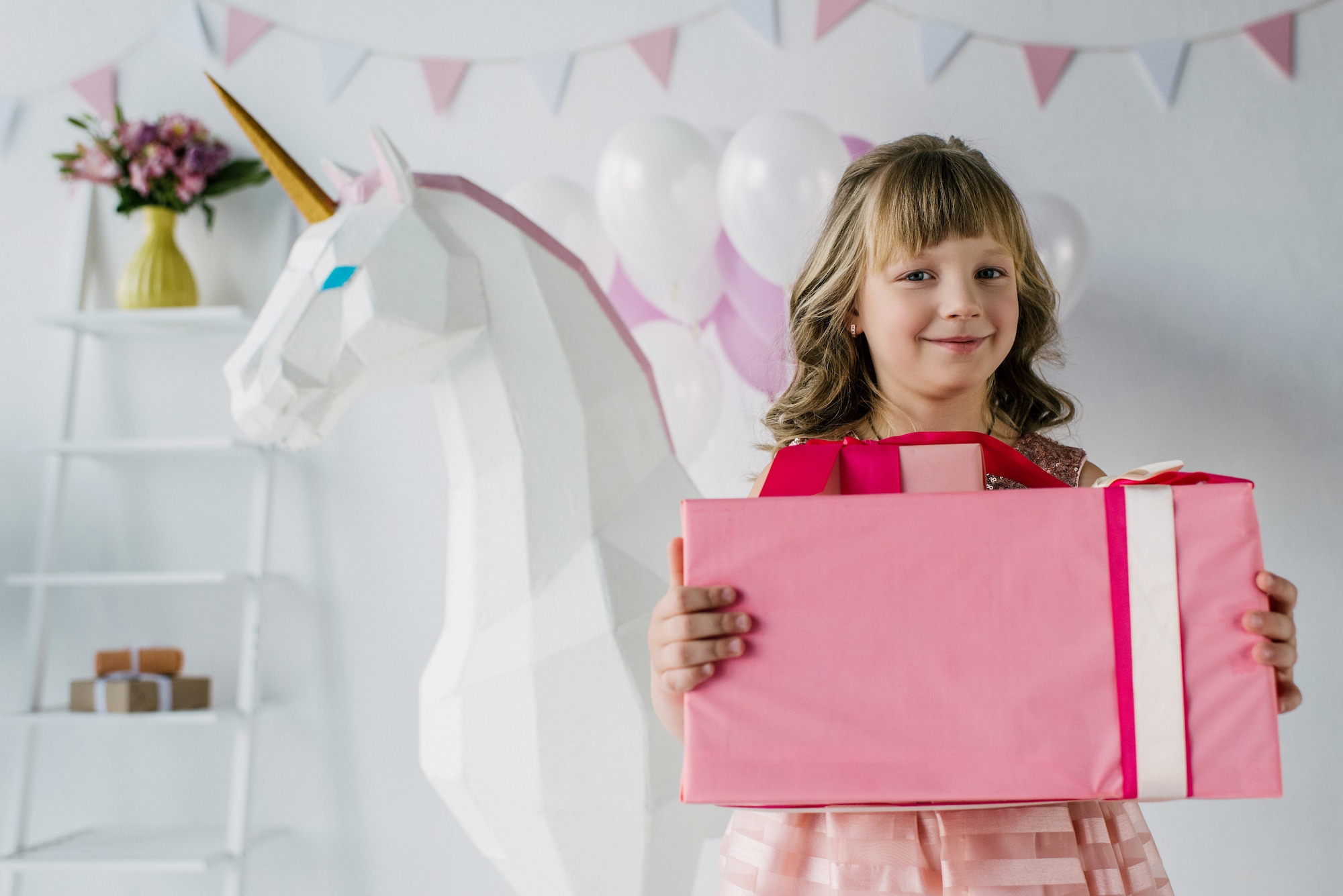 Mädchen steht mit rosa Geschenk vor einem weißen Einhorn in einem Zimmer mit rosa und weißen Wimpeln. Beschriftung lautet: Pferdemädchen und Pferdejungs freuen sich selbstverständlich über alle Geschenkideen rund um Pferde.