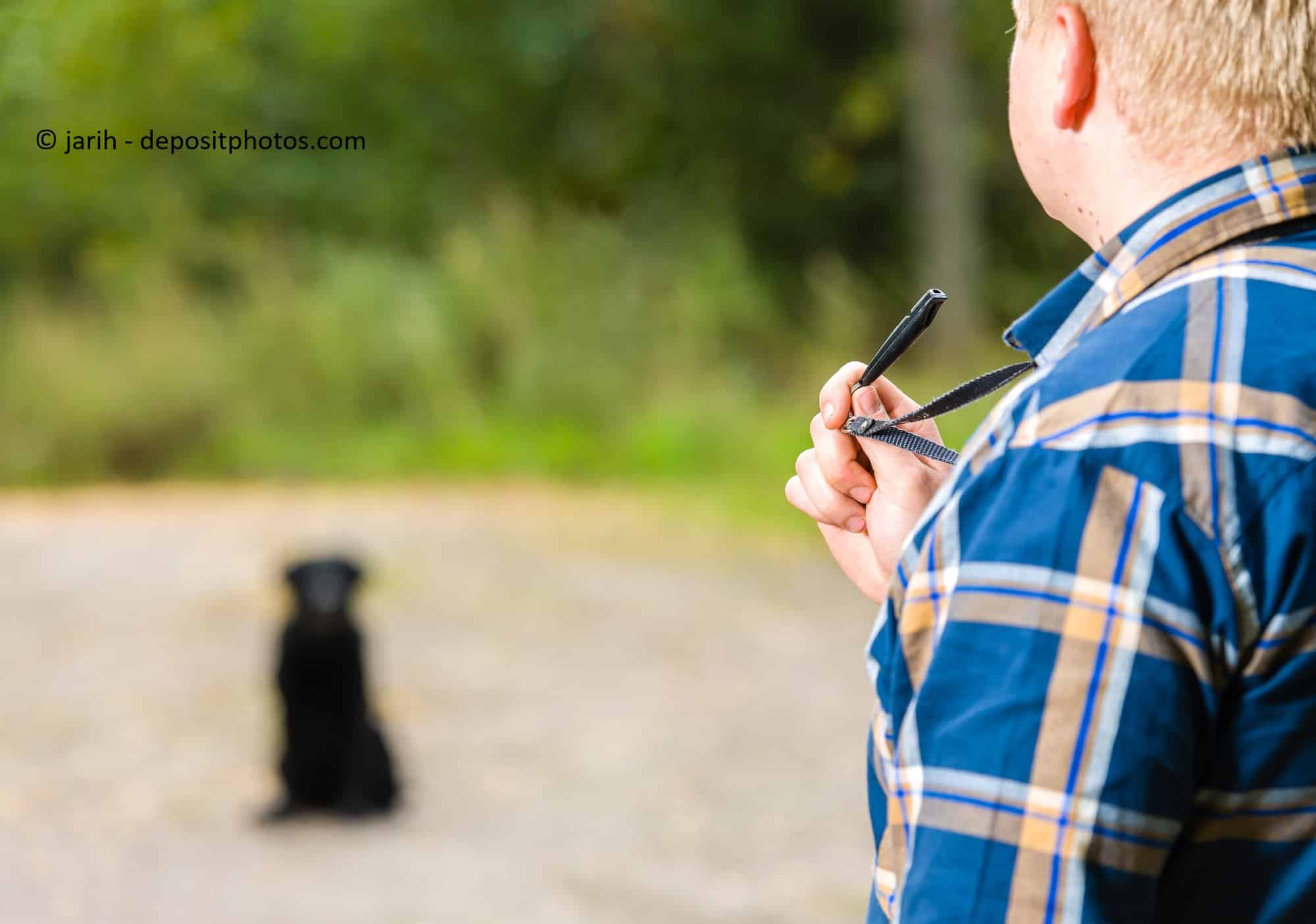 Ein Mann hält eine Hundepfeife in der Hand. Vor ihm sitzt sein Hund.